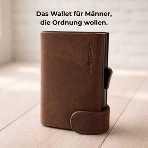 Könnte beinhalten: Ein braunes Lederportemonnaie mit schwarzem Akzent und Verschluss. Das Portemonnaie trägt den Text "G-WALLRET" vertikal auf der Seite. Der Text "Das Wallet für Männer, die Ordnung wollen" ist oben aufgedruckt.
