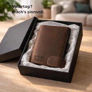 Könnte beinhalten: Ein braunes Lederportemonnaie mit dem Text "Bester Papa" wird in einer offenen schwarzen Geschenkbox mit weißem Seidenpapier präsentiert. Das Portemonnaie hat einen Magnetverschluss und einen Metallakzent. Der Text "Vaterstag? Mach's sinnvoll" ist sichtbar.