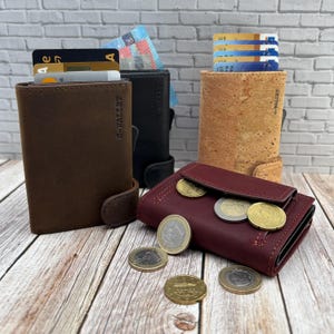 Könnte beinhalten: Eine Auswahl an Geldbörsen in verschiedenen Farben und Materialien, darunter braunes Leder, schwarzes Leder und Kork. Die Geldbörsen werden mit Kreditkarten und Münzen gezeigt, um ihre Speicherkapazität und ihr Design hervorzuheben. Die Geldbörsen haben den Text "G-WALLET".