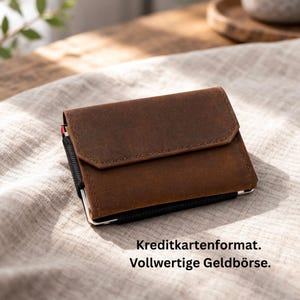 Könnte beinhalten: Ein braunes Lederportemonnaie mit Klappenverschluss. Das Portemonnaie hat ein schwarzes Gummiband und einen silberfarbenen Metallrahmen. Der Text "Kreditkartenformat. Vollwertige Geldbörse" ist unten im Bild zu sehen.