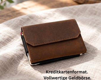 May include: A brown leather wallet with a flap closure. The wallet has a black elastic band and a silver-colored metal frame. The text "Kreditkartenformat. Vollwertige Geldbörse" is visible at the bottom of the image.