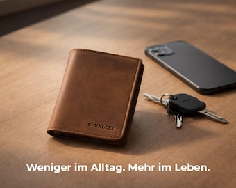 Op de afbeelding: Een bruine leren portemonnee met de tekst "G-WALLET" ligt op een houten oppervlak, naast een smartphone en een sleutelbos. De portemonnee is rechthoekig en gesloten. De tekst "Weniger im Alltag. Mehr im Leben." is ook aanwezig.