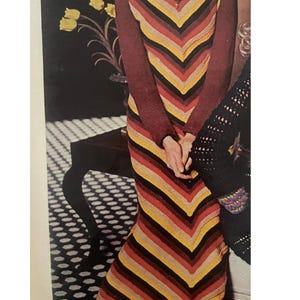 May include: A vintage-style, long-sleeved dress with a chevron pattern in shades of red, yellow, and brown. The dress has a fringed hem and a decorative element at the neckline. The sleeves are a solid burgundy color.