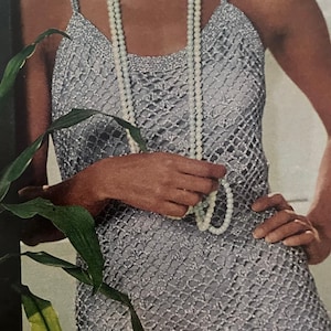 Könnte beinhalten: Ein silbernes Häkeltop mit Spaghettiträgern. Das Top hat ein netzartiges Muster und wird mit einer langen Kette aus weißen Perlen kombiniert. Das Bild ist eine Nahaufnahme, die die Textur des Stoffes und des Schmucks zeigt.