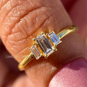 May include: A gold ring featuring three rectangular, clear gemstones. The ring's band is thin and gold-colored. The gemstones are set in a row, with the center stone slightly larger than the side stones. The ring is shown on a finger.
