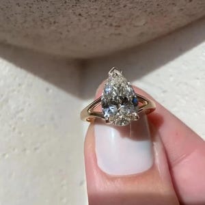 May include: A gold ring featuring a large, pear-shaped diamond. The diamond is clear with multiple facets, and the ring has a simple, elegant band. The ring is held up by a person.