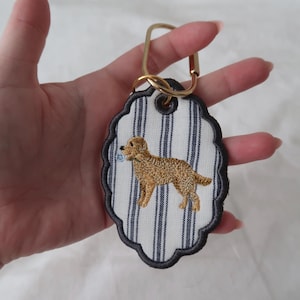 May include: An oval-shaped keyring with a scalloped edge featuring a white fabric background with vertical navy blue stripes. An embroidered golden-coloured dog is centred on the fabric, and the keyring includes a gold-toned metal clasp.