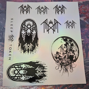May include: A sticker sheet featuring black Sleep Token band logos and graphic designs on a holographic background. The sheet includes multiple variations of the logo, a moon-like sphere with tentacles, and two arched, dripping architectural motifs.