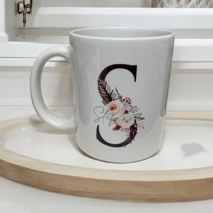 May include: White ceramic mug with a black letter "S" decorated with floral and feather designs. The name "Stefanie" is written in cursive below the letter.