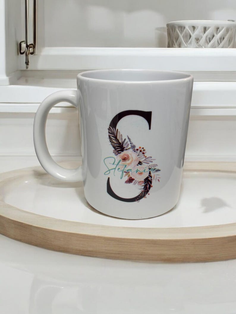 May include: White ceramic mug with a black letter "S" decorated with floral and feather designs. The name "Stefanie" is written in cursive below the letter.
