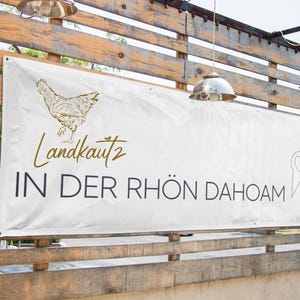 May include: White banner with a gold chicken illustration and the text "Landkautz 2 IN DER RH&Ouml;N DAHOAM" in black. A gold pin with a heart is in the bottom right corner.