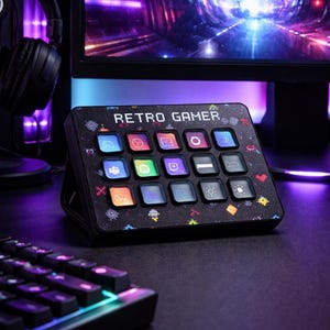 Könnte beinhalten: Ein schwarzes "Retro Gamer" Bedienfeld mit mehreren beleuchteten Tasten. Das Panel hat ein pixeliges Design mit verschiedenen Symbolen. Eine Tastatur und Kopfhörer sind ebenfalls im Hintergrund zu sehen, was auf ein Gaming-Setup hindeutet.