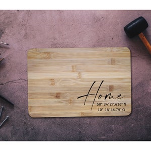 Könnte beinhalten: Ein Schneidebrett aus Holz mit dem Wort "Home" und Koordinaten darauf. Das Brett ist von Werkzeugen umgeben, darunter ein Hammer, eine Zange und Nägel.