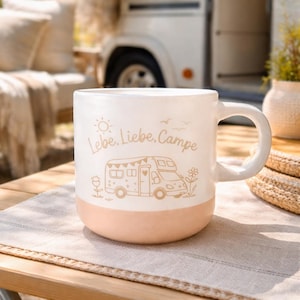 Könnte beinhalten: Weißer Keramikbecher mit pfirsichfarbenem Boden, mit einer Wohnmobil-Illustration und den Worten "Lebe, Liebe, Campe". Der Becher hat einen großen Henkel und steht auf einem Holztisch mit einem neutralen Platzset. Im Hintergrund ist ein Wohnmobil zu sehen.