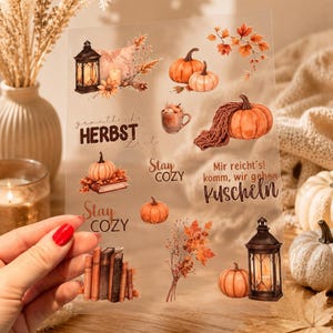 Könnte beinhalten: Ein transparenter Stickerbogen mit herbstlichen Illustrationen wie orangefarbenen Kürbissen, einer schwarzen Laterne, einer Tasse, einem Bücherstapel und Herbstlaub. Text: "gemütliche HERBST Zeit", "Stay COZY" und "Mir reicht's! komm, wir gehen Kuscheln".