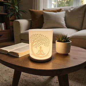 Könnte beinhalten: Eine zylindrische, cremefarbene Lampe mit einem Baum- und Meditationsfigurendesign. Die Lampe steht auf einem dunkelbraunen Sockel und auf einem runden Holztisch. Ein kleiner Topf mit Sukkulenten und ein offenes Buch befinden sich ebenfalls auf dem Tisch.