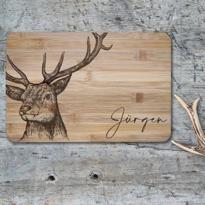 May include: A rectangular bamboo cutting board featuring a dark brown laser-engraved illustration of a stag head with large antlers on the left side and the name J&uuml;rgen written in a cursive script font on the right side of the board.