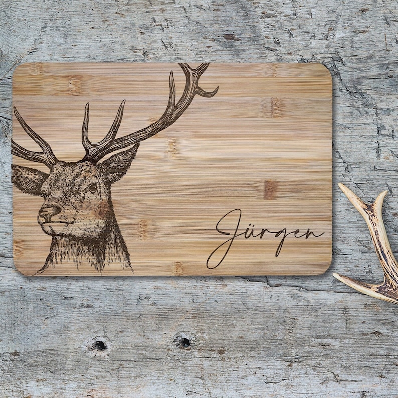 May include: A rectangular bamboo cutting board featuring a dark brown laser-engraved illustration of a stag head with large antlers on the left side and the name J&uuml;rgen written in a cursive script font on the right side of the board.