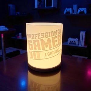 Könnte beinhalten: Eine zylindrische Lampe mit den Worten "PROFESSIONAL GAMER LOADING..." auf der Seite eingraviert. Die Lampe strahlt ein warmes, weiches Licht aus und steht auf einem dunklen Holztisch. Der Hintergrund zeigt Spielkonsolen und Controller.