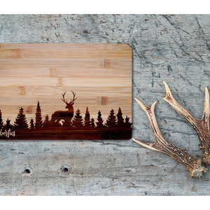 May include: A brown bamboo cutting board with a laser-etched design of a deer standing in a forest. The text "Hubertus" is engraved on the board.
