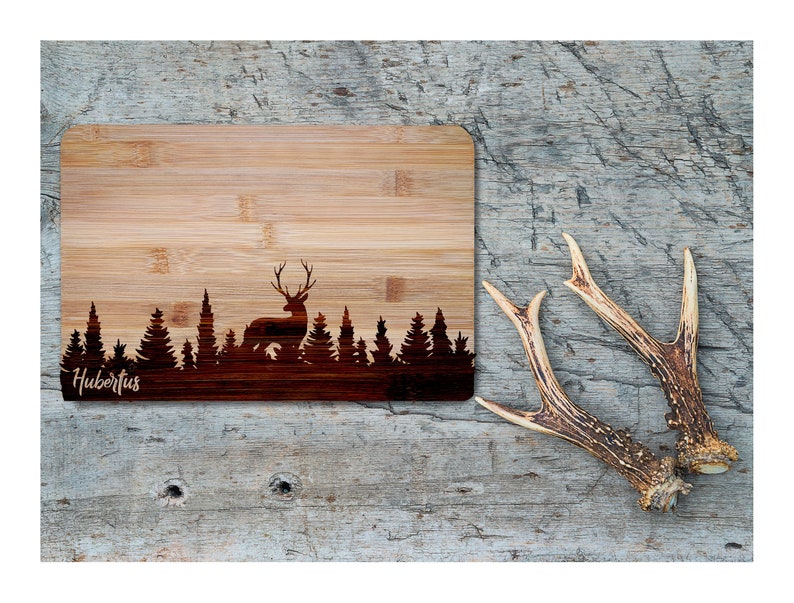 May include: A brown bamboo cutting board with a laser-etched design of a deer standing in a forest. The text "Hubertus" is engraved on the board.