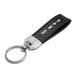 May include: Black and silver key chain with the text "XT4" and a Cadillac logo. The key chain has a carbon fiber pattern on the strap and a silver metal clasp and key ring.