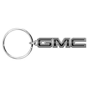 May include: A silver-colored keychain with a large ring and a black and silver GMC logo. The logo is a stylized text design with a black background and silver outline. The keychain is likely made of metal.