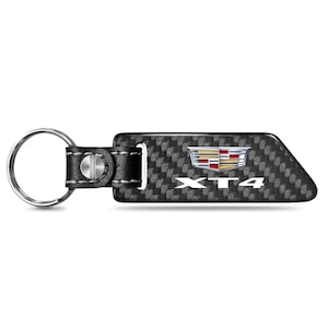 May include: A black and gray carbon fiber keychain with a silver key ring and black leather strap. The keychain features the Cadillac logo and the letters "XT4" in white.
