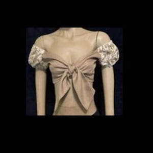 May include: A beige cropped top featuring a front tie detail and off-the-shoulder puff sleeves. The sleeves are adorned with a white floral pattern, while the bodice is a solid beige colour. The garment has a sweetheart neckline and a cropped hem.