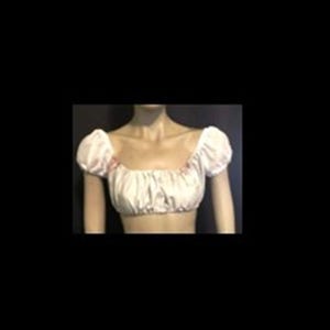 May include: A white off-the-shoulder crop top featuring short puff sleeves with gathered elastic detailing at the shoulders and neckline. The garment has a straight hemline and a soft, lightweight appearance with subtle ruching across the front bodice.