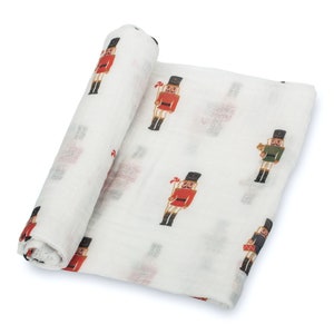 May include: White muslin swaddle blanket with a repeating pattern of colorful toy soldier illustrations. The toy soldiers are depicted in red, green, and blue uniforms with black hats. The blanket is rolled up, showing the pattern.