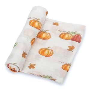 May include: A rolled-up, white muslin swaddle blanket with a repeating pattern of orange pumpkins and brown autumn leaves. The pumpkins have green vines. The blanket is made of a soft, lightweight fabric.