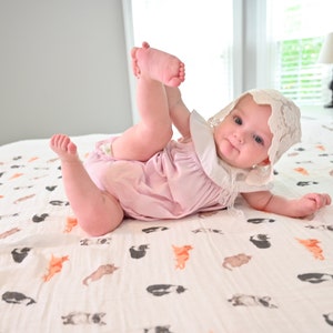 May include: A baby wearing a pink romper and a white lace bonnet lies on a bed. The baby is playfully holding their foot. The bedspread features a pattern of cats in various colors. The room has a window and a lamp.