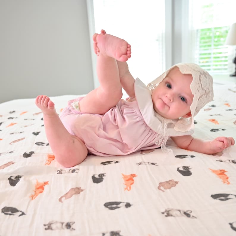 May include: A baby wearing a pink romper and a white lace bonnet lies on a bed. The baby is playfully holding their foot. The bedspread features a pattern of cats in various colors. The room has a window and a lamp.
