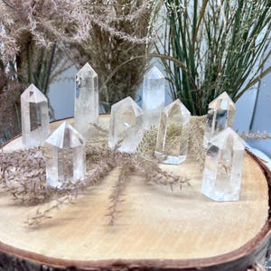 Könnte beinhalten: Eine Sammlung von acht klaren Quarzkristallspitzen, die auf einer Holzoberfläche angeordnet sind. Die Kristalle sind in verschiedenen Größen und Formen, wobei einige eine spitzere Spitze haben als andere.