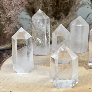 Könnte beinhalten: Eine Sammlung klarer Quarz-Kristallspitzen mit facettierten Seiten und spitzen Spitzen. Die Kristalle sind auf einer hellbraunen Holzoberfläche angeordnet, mit getrockneten Pflanzen im Hintergrund. Die Kristalle variieren in Größe und Form.