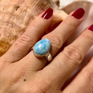 Könnte beinhalten: Ein Silberring mit einem tropfenförmigen, hellblauen Larimar-Edelstein. Der Stein hat ein wolkiges, marmoriertes Aussehen. Der Ring wird an einer Hand mit rotem Nagellack gezeigt.