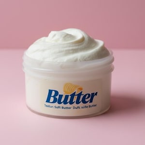 May include: A small, clear plastic container filled with white, whipped butter. The container has a label with the word "Butter" in blue, a yellow butter curl graphic, and text that reads "Textur: Soft Butter Duft: süße Butter". The background is pink.