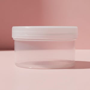 May include: A clear, cylindrical plastic container with a matching translucent lid. The container features a smooth, wide-mouth design with a screw-top closure, providing a simple and functional storage solution for various small items.
