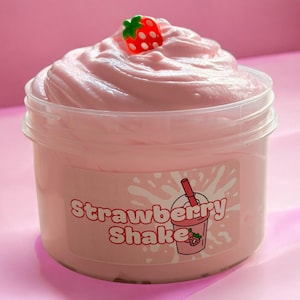 May include: A clear plastic container filled with pink slime, topped with a swirl and a small strawberry charm. The container has a label that reads "Strawberry Shake" with a graphic of a milkshake.