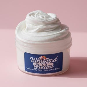 May include: A jar of white whipped cream with a swirl design on top. The jar has a blue label with the words "Whipped Cream" and a strawberry illustration. The text below the words reads "Textur: Fluffy Whip - Duft: Vanille Sahne".