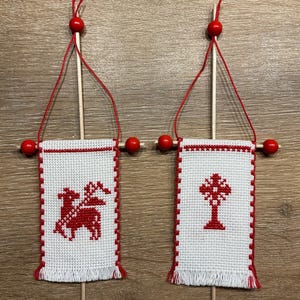 Könnte beinhalten: Zwei kleine, weiß gekreuzte Banner mit roten Akzenten. Ein Banner zeigt ein rotes Pferd und einen Reiter, das andere ein rotes Kreuz. Jedes Banner ist mit Holzdübeln mit roten Perlen und Schnur befestigt, wodurch ein dekoratives Hängeornament entsteht.