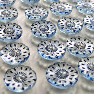 May include: A collection of clear glass buttons with a floral design. Each button features a central faceted gem surrounded by a pattern of blue and white detailing. The buttons have a scalloped edge and are arranged in a grid.