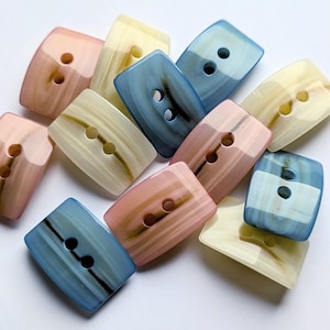 Könnte beinhalten: Eine Ansammlung rechteckiger Knöpfe in verschiedenen Farben, darunter Rosa, Blau und Creme. Jeder Knopf hat zwei Löcher und ein marmoriertes Aussehen. Die Knöpfe sind in einem verstreuten Muster angeordnet.