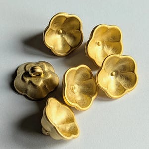 Könnte beinhalten: Sechs goldfarbene, blütenförmige Knöpfe. Jeder Knopf hat ein detailliertes Blütenblattdesign und ein kleines Loch in der Mitte. Die Knöpfe sind auf einer hellgrauen Oberfläche angeordnet.