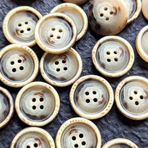 Può includere: Una collezione di 12 bottoni beige e marroni con quattro fori ciascuno. I bottoni hanno un motivo screziato e un bordo rialzato.