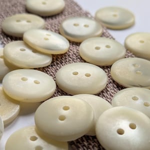 Op de afbeelding: Een verzameling witte parelmoerachtige knopen met elk twee gaten. De knopen zijn gerangschikt op een bruin stofoppervlak.