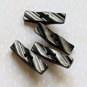 May include: Five dark grey and silver rectangular buttons with two holes each. The buttons have a diagonal striped pattern and a slightly curved shape. The buttons are arranged on a light background.