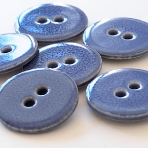 Puede incluir: Primer plano de varios botones de cerámica azules con dos agujeros cada uno. Los botones tienen un acabado brillante y una superficie texturizada. Los botones son redondos y tienen un borde ligeramente elevado.