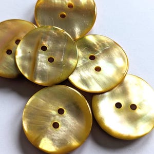 Può includere: Una collezione di bottoni rotondi, iridescenti, giallo dorato, ciascuno con due fori. I bottoni hanno una lucentezza madreperlacea e sono disposti in modo leggermente sovrapposto, evidenziando la loro consistenza naturale. Ideale per cucito e progetti artigianali.